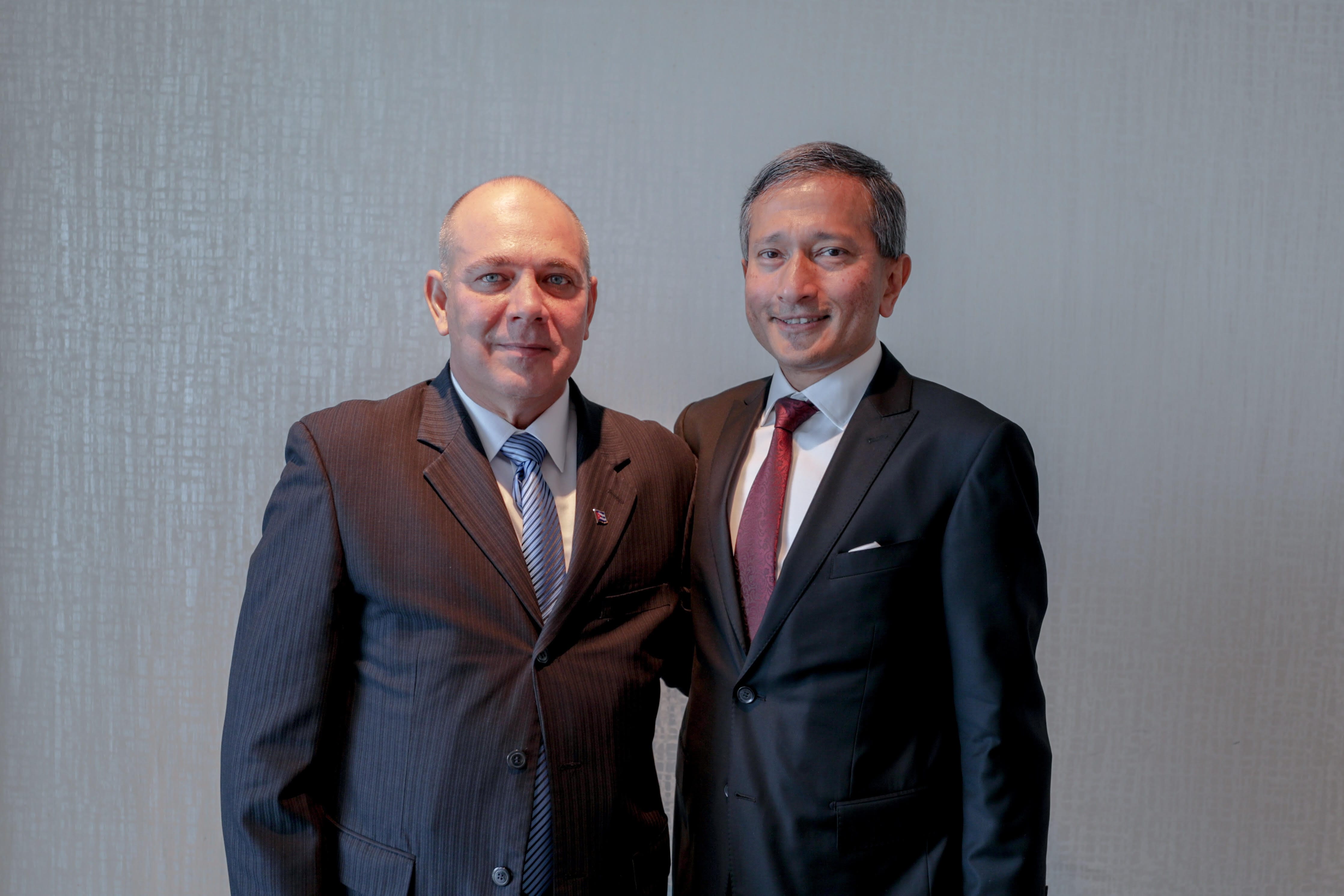 Two men in suits stand side-by-side against a textured, light gray wall.